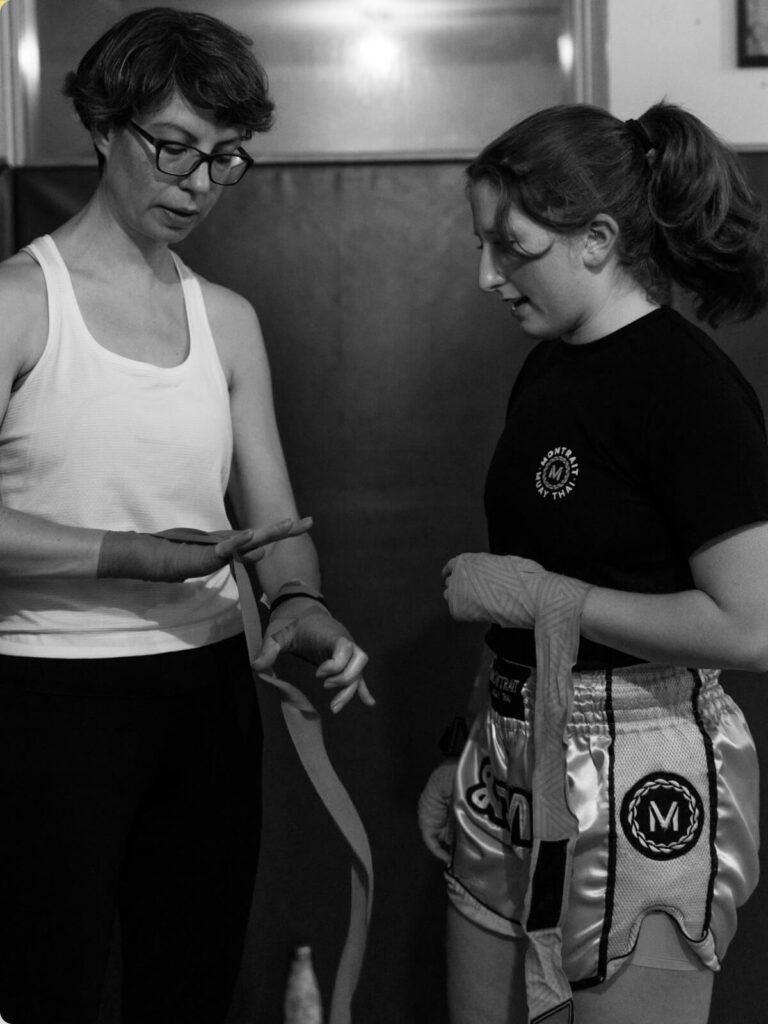 An experienced Muay Thai student helps a beginner prepare for training by wrapping their hands before class at Montrait Muay Thai Gym.